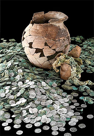 Gesamtansicht des Münzschatzes. Auf dem Münzhaufen liegen im Kontrast zu den korrodierten, nicht restaurierten Münzen, die silberglänzenden restaurierten Münzen. In der Mitte zwischen den Münzen steht das Münzgefäß ein einfacher Keramiktopf. Rechts daneben die Becherkachel mit erhaltenen Textilien. Vorne rechts liegt der kleine Bronzering.